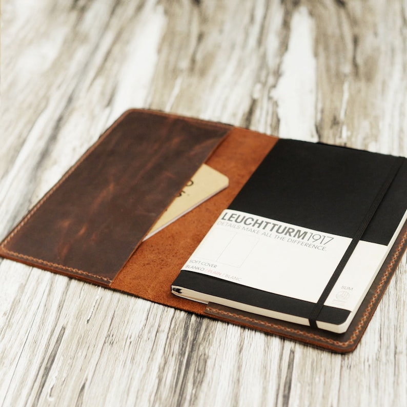 May include: A brown leather notebook cover with a black notebook inside. The notebook has a black elastic band and a white label that says "LEUCHTTURM1917 DETAILS MAKE ALL THE DIFFERENCE."