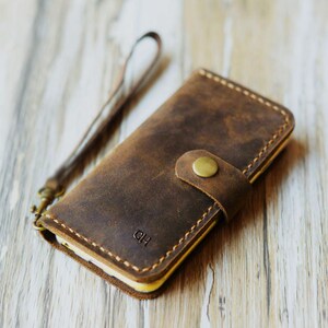 Peut inclure: Une coque de t&eacute;l&eacute;phone en cuir marron avec une sangle et une fermeture &agrave; bouton dor&eacute;e. La coque porte les initiales "DH" grav&eacute;es sur le devant.