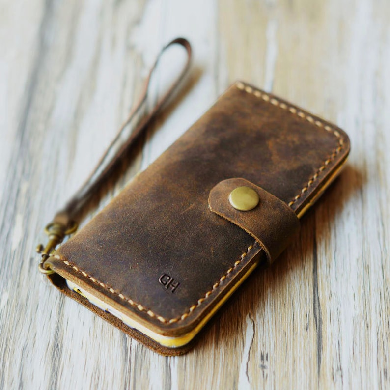 Peut inclure: Une coque de t&eacute;l&eacute;phone en cuir marron avec une sangle et une fermeture &agrave; bouton dor&eacute;e. La coque porte les initiales "DH" grav&eacute;es sur le devant.