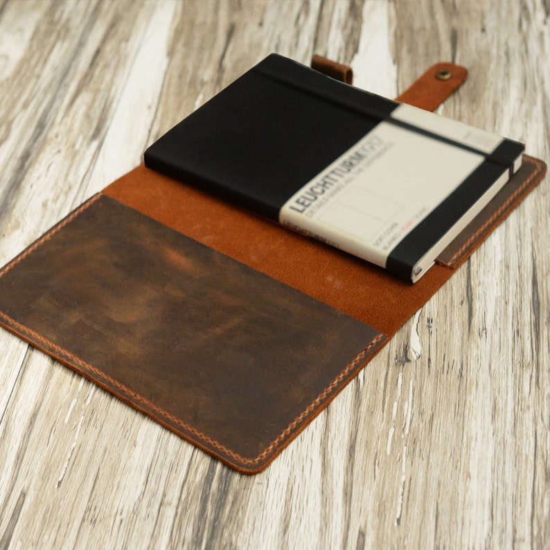 May include: A brown leather notebook cover with a black notebook inside. The cover has a distressed look and is stitched around the edges. The notebook has a black elastic band and the text "LEUCHTTURM1917" printed on the cover.
