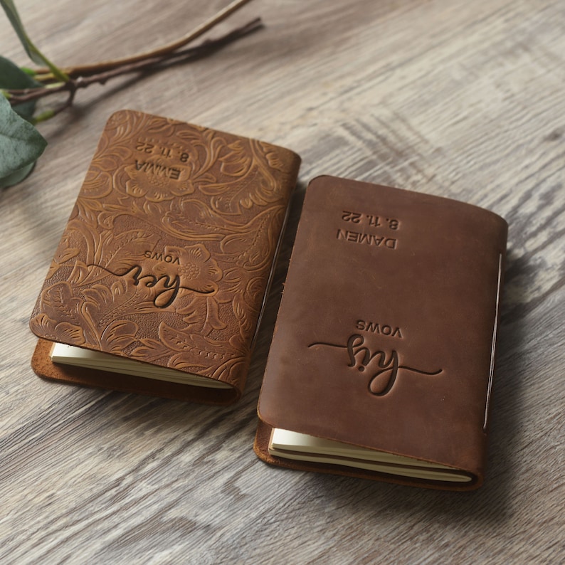 May include: Two brown leather journals with embossed floral designs. The covers are personalized with the text "Our Vows" and the dates "8.11.22" and "8.11.22".