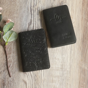 May include: Two black leather vow books with embossed floral designs. The book on the left says "her vows" and the book on the right says "his vows".