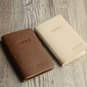 May include: Two leather-bound vow books, one brown and one beige, with the word "VOWS" embossed on the front. The date "08.04.22" is embossed on the brown book and "06.04.22" is embossed on the beige book.