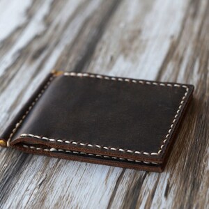 May include: A brown leather wallet with a money clip and white stitching. The wallet is lying on a light brown wooden surface.