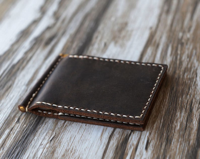 May include: A brown leather wallet with a money clip and white stitching. The wallet is lying on a light brown wooden surface.