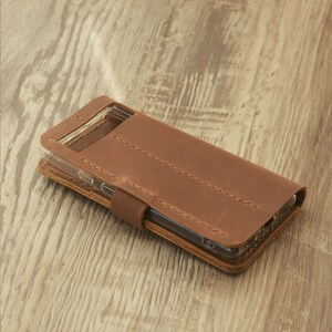 Peut inclure: Une coque de t&eacute;l&eacute;phone en cuir marron avec un insert en plastique transparent pour le t&eacute;l&eacute;phone. La coque pr&eacute;sente des d&eacute;tails de couture et un rabat qui se replie sur le t&eacute;l&eacute;phone.