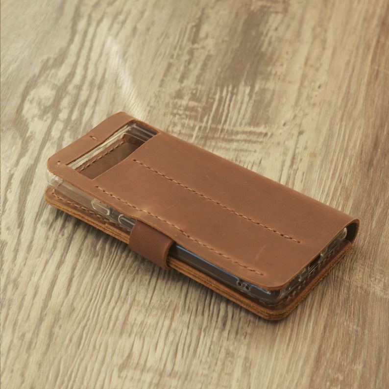 Peut inclure: Une coque de t&eacute;l&eacute;phone en cuir marron avec un insert en plastique transparent pour le t&eacute;l&eacute;phone. La coque pr&eacute;sente des d&eacute;tails de couture et un rabat qui se replie sur le t&eacute;l&eacute;phone.