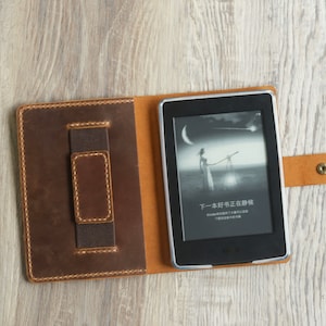 Op de afbeelding: Een bruine leren hoes met een ingebouwde elastiekband om een e-reader vast te houden. De e-reader is open op een pagina met een zwart-wit afbeelding van een vrouw die in een veld staat met een ster aan de hemel. De tekst op het scherm luidt "下一本好书正在加载".