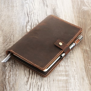May include: A brown leather notebook with a pen tucked into the elastic strap. The notebook has a white ribbon bookmark and is closed.