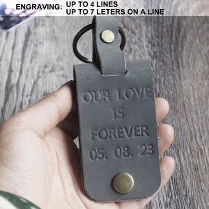 May include: A gray leather keychain with a metal ring and a brass button. The keychain is engraved with the text "OUR LOVE IS FOREVER 05.08.23".