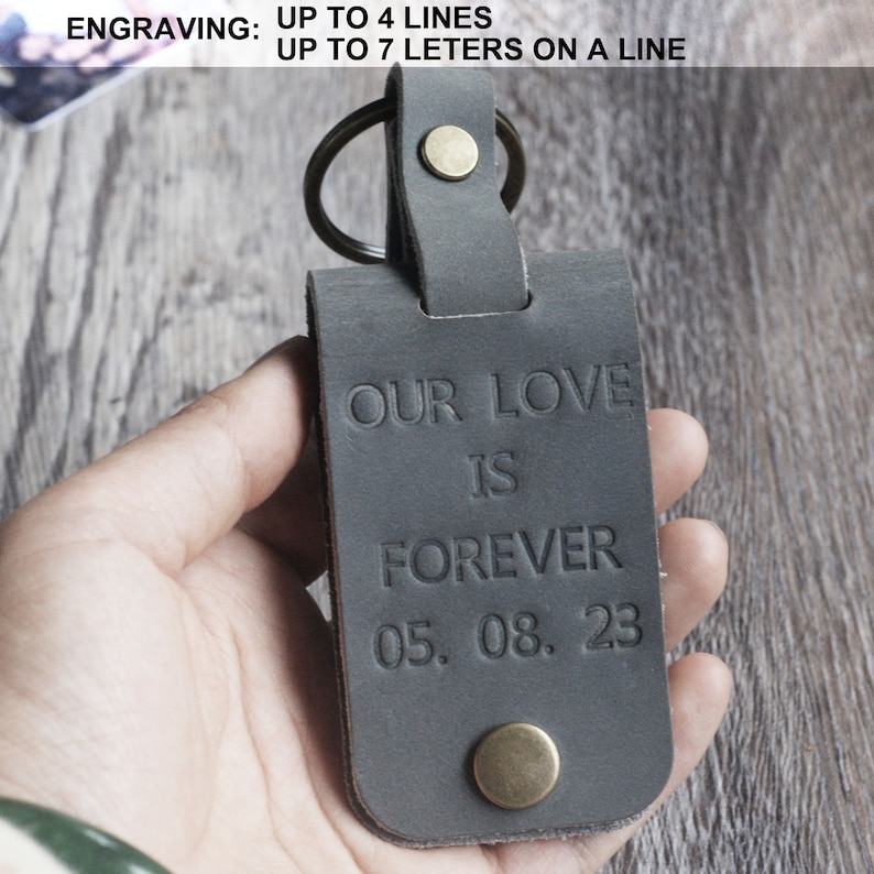 May include: A gray leather keychain with a metal ring and a brass button. The keychain is engraved with the text "OUR LOVE IS FOREVER 05.08.23".