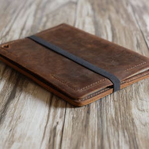 May include: A brown leather tablet case with a black elastic band closure. The case is lying on a wooden surface.