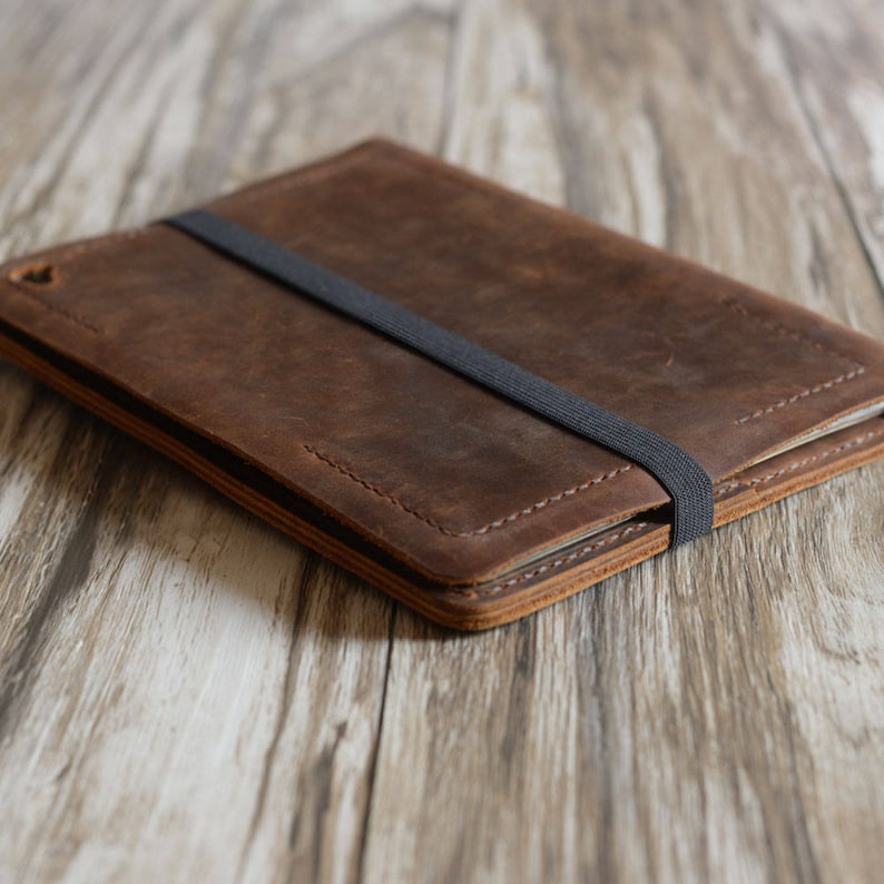 May include: A brown leather tablet case with a black elastic band closure. The case is lying on a wooden surface.