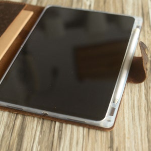 May include: A brown leather case with a flap that folds over to protect a tablet and stylus. The tablet is black and the stylus is white.