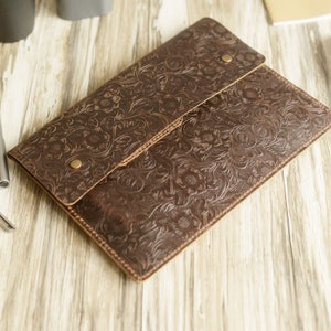 Peut inclure: Une pochette pour ordinateur portable en cuir marron avec un motif en relief floral. La pochette a une fermeture &agrave; rabat avec un bouton pression.