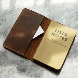 May include: A brown leather notebook cover with a Field Notes memo book inside. The cover has a pocket on the front and is open to reveal the notebook. The notebook is a 43-page memo book made in the USA.