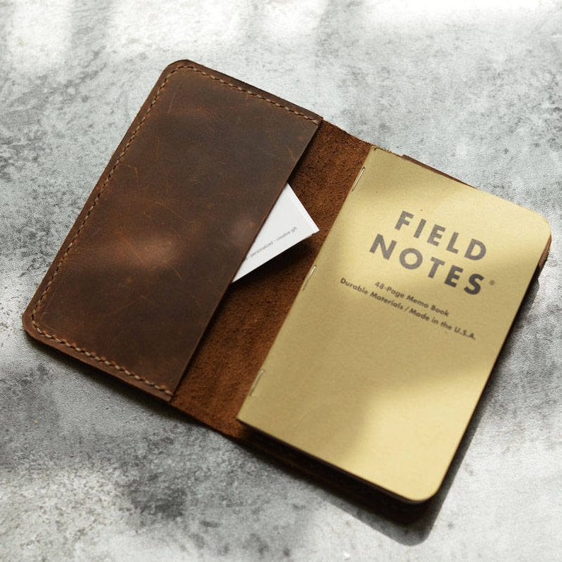 May include: A brown leather notebook cover with a Field Notes memo book inside. The cover has a pocket on the front and is open to reveal the notebook. The notebook is a 43-page memo book made in the USA.
