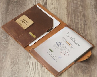 Op de afbeelding: Een bruine leren portfolio met een klep sluiting en een vak voor een notitieblok. De portfolio heeft een transparante plastic hoes voor het bewaren van papieren. De tekst "Project Milestones" staat op een vel papier in de portfolio.