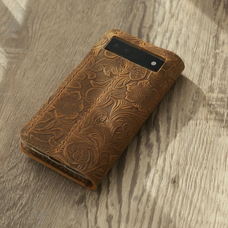 May include: A brown leather phone case with a floral embossed design. The case has a cutout for the phone's camera lens.