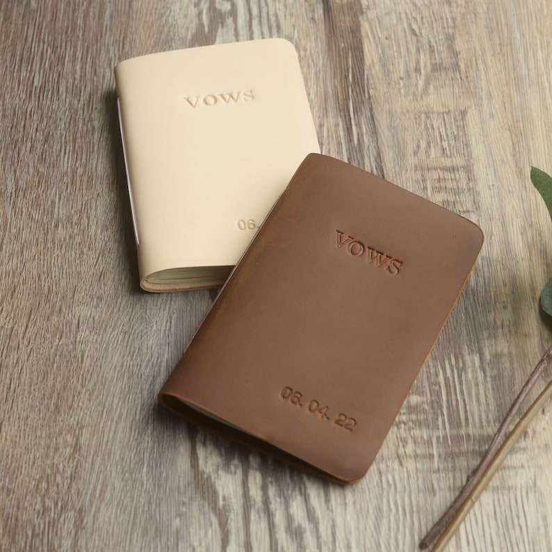 May include: Two leather-bound vow books, one brown and one cream, with the word "Vows" embossed on the front. The brown book has the date "05.04.22" embossed on the front.