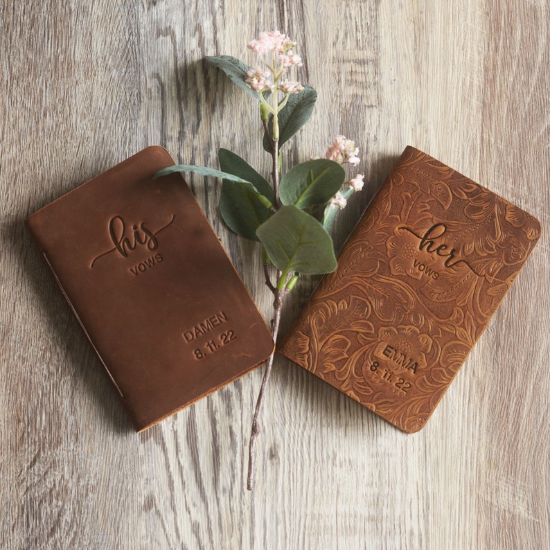 May include: Two brown leather vow books with the words "his vows" and "her vows" engraved on the front. The book on the left has the name "Damen" and the date "8.11.22" engraved on the front. The book on the right has the name "Emma" and the date "8.11.22" engraved on the front.