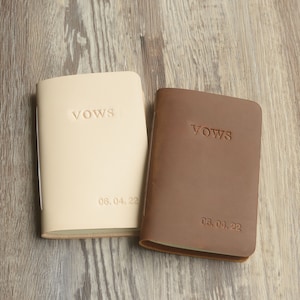 May include: Two leather-bound vow books with the word "Vows" embossed on the front. The books are beige and brown and have the date "06.04.22" printed on the bottom.