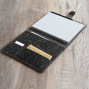 May include: A black leather portfolio with an embossed floral design. The portfolio is open, revealing a white tablet with a black stylus and a brown card holder.