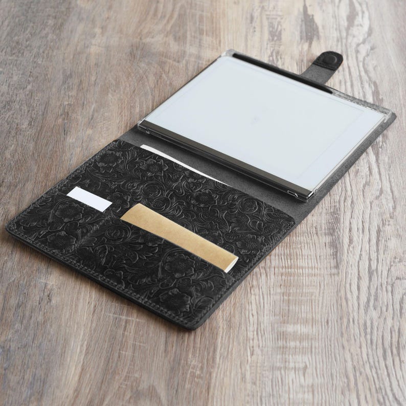 May include: A black leather portfolio with an embossed floral design. The portfolio is open, revealing a white tablet with a black stylus and a brown card holder.