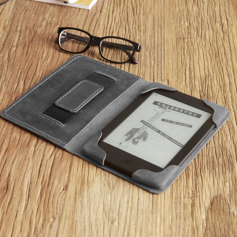 Puede incluir: Una funda de cuero gris para lector electr&oacute;nico con una correa el&aacute;stica integrada para sujetar el lector electr&oacute;nico en su lugar. La funda est&aacute; abierta y se puede ver un lector electr&oacute;nico con una pantalla que muestra texto en chino.