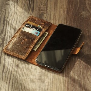 May include: A brown leather phone case with a floral embossed design. The case has slots for credit cards and cash. A black smartphone is inside the case.