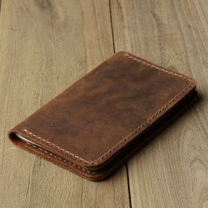 May include: A brown leather notebook cover with white stitching. The cover is closed and lying on a wooden surface.