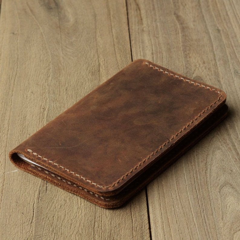 May include: A brown leather notebook cover with white stitching. The cover is closed and lying on a wooden surface.