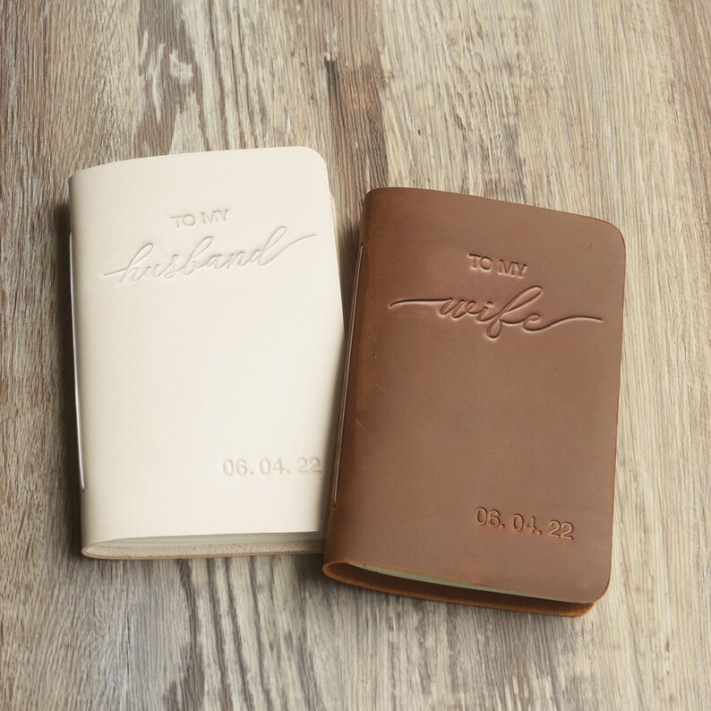 May include: Two leather-bound journals, one white and one brown, with the words "To My Husband" and "To My Wife" embossed on the covers. The date "06.04.22" is also embossed on each journal.