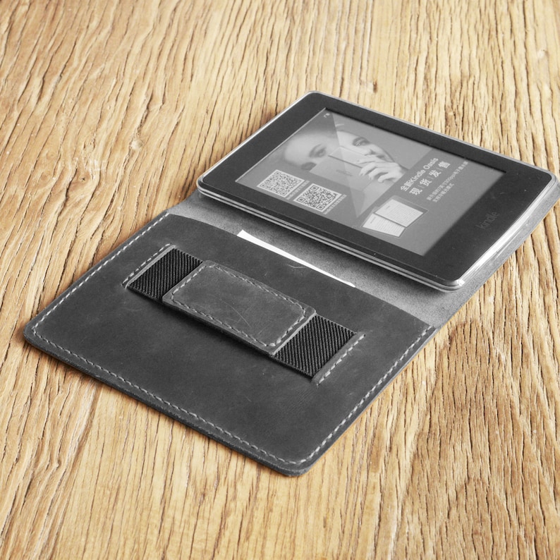 May include: A black leather e-reader case with a built-in elastic strap to hold the e-reader in place. The case is open and an e-reader is inside.