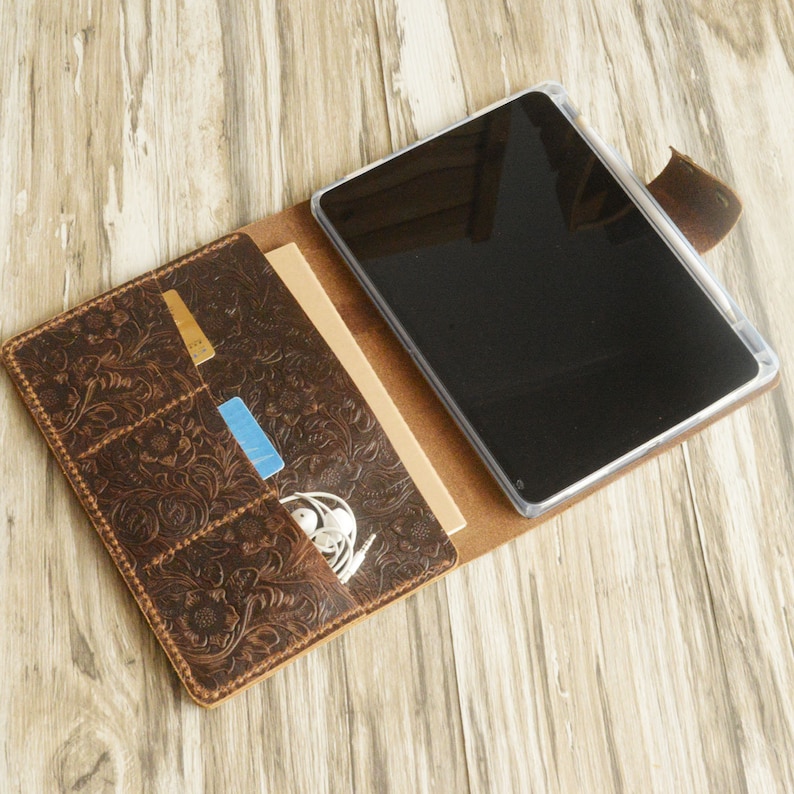 May include: A brown leather folio case with an embossed floral design. The case has multiple pockets for cards and other small items. A tablet is visible inside the case.