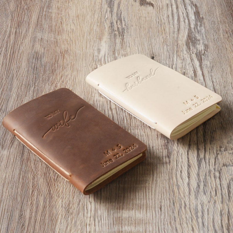 May include: Two leather-bound journals with personalized inscriptions. One is brown with the inscription "To my wife" and the other is cream with the inscription "To my husband". Both journals have the date "June 22, 2024" and the initials "M+S" embossed on the cover.