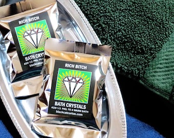 May include: Two silver foil packets of bath crystals labeled RICH BITCH with a diamond graphic. The text reads: ADD 1/2 PKG. TO A WARM BATH blackcatcurios.com. The packets are resting on a silver oval tray next to a dark green textured fabric.