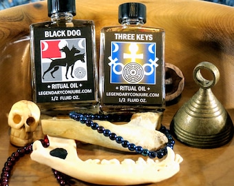 Puede incluir: Dos botellas de vidrio de aceite ritual con tapas negras, etiquetadas como "BLACK DOG" y "THREE KEYS". Las botellas están acompañadas de una calavera, un hueso, un collar de cuentas y una campana de latón. Las etiquetas incluyen el texto "+ RITUAL OIL + LEGENDARYCONJURE.COM 1/2 FLUID OZ."