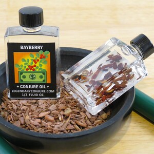 May include: Two clear glass bottles with black caps containing a red and green liquid. The bottles are labeled "Bayberry + Conjure Oil + LEGENDARYCONJURE.COM 1/2 FLUID OZ.". The bottles are sitting on a brown surface.