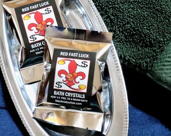 May include: Two silver foil packets of Red Fast Luck bath crystals featuring a red fleur-de-lis and dice graphic with dollar signs. The labels include the text: RED FAST LUCK, BATH CRYSTALS, ADD 1/2 PKG. TO A WARM BATH, and blackcatcurios.com.