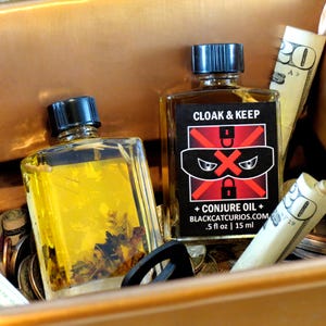May include: Two glass bottles with black caps, one labeled "CLOAK & KEEP" with a graphic of a masked face and the text "+ CONJURE OIL + BLACKCATCURIOS.COM .5 fl oz | 15 ml". The other bottle contains a yellow liquid with plant matter. The bottles are in a copper-colored container with money and coins.