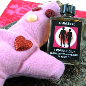 May include: A small bottle of brown liquid with a black label that reads "Adam & Eve + Conjure Oil + LegendaryConjure.com 1/2 Fluid Oz.". The label also features a red circle with a silhouette of two nude figures standing side-by-side. The bottle is sitting on a silver tray with a pink fabric heart-shaped object with red glitter.