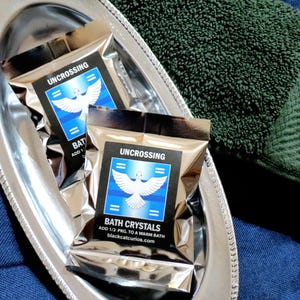 May include: Two silver metallic packets of Uncrossing bath crystals featuring a white dove graphic on a blue background. The text reads Uncrossing, Bath Crystals, Add 1/2 pkg. to a warm bath, and blackcatcurios.com. The packets rest in a silver tray.