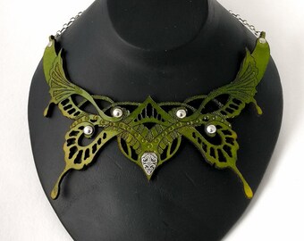 Puede incluir: Un collar con forma de mariposa verde que presenta intrincados patrones recortados, tachuelas de metal plateado y un acento central de filigrana plateada. La pieza está unida a una fina cadena de metal plateado en ambos lados.