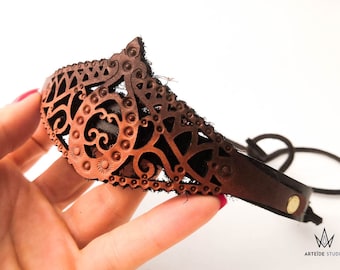 May include: A brown leather headband featuring an intricate laser-cut filigree pattern with small circular embossed details. The accessory has a pointed center section, a smooth leather strap, and a metallic snap fastener. The Arteide Studio logo is visible.
