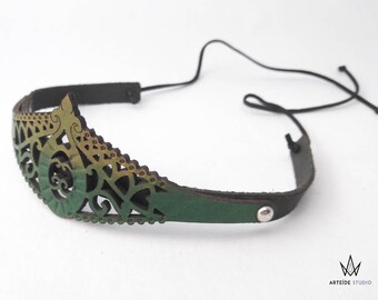 May include: A leather headband featuring a laser-cut filigree design in a gradient of green and gold. The piece includes a dark brown leather strap secured with a silver-toned metal rivet and thin black cords for tying at the back of the head.