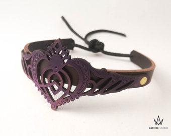 May include: A dark brown leather bracelet featuring a purple-toned, laser-cut heart-shaped centerpiece with intricate filigree details. The band is secured with a gold-toned metal rivet and a black cord tie. The logo ARTEIDE STUDIO is visible.