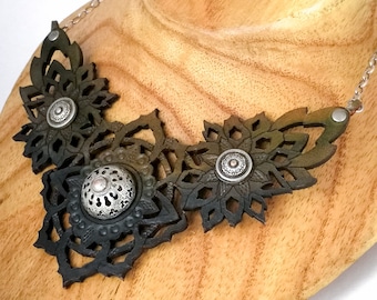 Puede incluir: Un collar llamativo con formas florales de cuero cortadas con láser en tonos verde oscuro y negro, dispuestas en capas. El diseño incluye tres adornos circulares de metal plateado con detalles de filigrana, unidos a una cadena de color plateado.