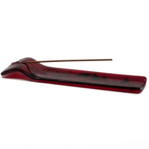 May include: A red glass incense holder with a long, rectangular shape and a curved end. A brown incense stick rests in the holder. The holder is on a white background.
