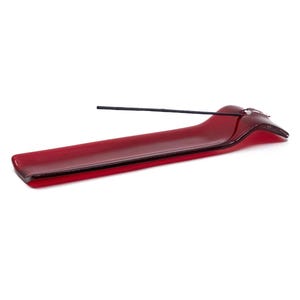 May include: A red glass incense holder with a sleek, modern design. The holder is rectangular with a curved end and a small hole to hold the incense stick. A black incense stick is placed in the holder. The background is white.